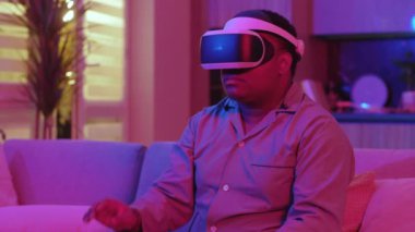 Sanal gerçeklik kulaklığı takan melez genç adam evde neon ışığı altında oturuyor. Eğlenceli oyundu. VR gözlükleri. Sosyal ağ robotu. Yavaş çekim