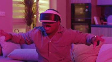 Evde, sanal gerçeklik kulaklığı takan genç bir adam kanepeye oturmuş korkmuş neon ışığı hissediyor. Eğlenceli oyundu. VR gözlükleri. Sosyal ağ robotu. Yavaş çekim