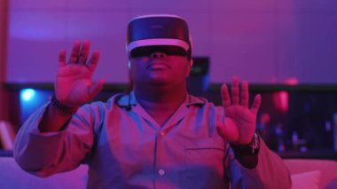 Sanal gerçeklik kulaklığı takmış gülümseyen melez genç adam evdeki ışıklı kanepede oturuyor. Eğlenceli oyundu. VR gözlükleri sosyal ağ robotu. Yavaş çekim