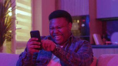 Gülümseyen genç Afrikalı Amerikalı adam mesaj yazıyor ve akıllı telefon ekranına bakıyor. Kanepede oturup evdeki sosyal ağlarda sohbet etmek. Yavaş çekim