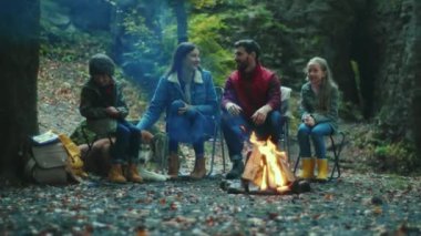 Komik aile sonbahar akşamı kamp ateşinin yanında oturur. Ebeveynleriyle birlikte çocuklar ormanda dinleniyorlar. Çocukluk ilişkisi kamp kuruyor. Kapatın. Yavaş çekim