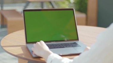 NEW YORK - 5 Eylül 2020: Kafenin yuvarlak ahşap masasında oturan ve yeşil ekranlı dizüstü bilgisayarla çalışan kadınların görüntüleri. Bilgisayarda yazan genç bir kadın. Güneşli bir gün. Yüzü olmadan. Kadın.