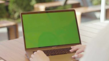 NEW YORK - 5 Eylül 2020: Yeşil ekranlı yeni gümüş dizüstü bilgisayarın dokunmatik pedini kullanan kadın elleri. Beyaz gömlekli bir iş kadınının kafein masasında oturup bilgisayarla çalışmasının, yazı yazmasının omuz manzarası.
