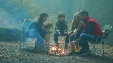 Aile, sonbahar akşamı ormanda kamp ateşinin etrafında oturur. Ormandaki bir yangında sosis kızartmak. Dostluk mutluluğu şömineyi pişirir. Kapatın. Yavaş çekim
