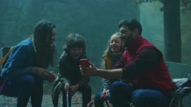 Köpekli bir aile, sonbahar akşamı ormanda kamp ateşinin etrafında oturur. Ormandaki bir yangında sosis kızartmak. Telefonu kullan. Arkadaşlık şömineyi pişirir. Kapatın. Yavaş çekim