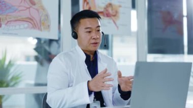 Asyalı pratisyen doktor kulaklık takıyor ve hastaya danışmanlık yapıyor. Uzaktan görüşme videosu, laptop konferansı, tıbbi randevu..