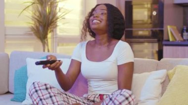 Geceleri, karanlık oturma odasındaki kanepede oturan Afrikalı Amerikalı kadın. Televizyon izlerken gülümseyen uzaktan kumandayla kanal değiştirmek. Yavaş çekim