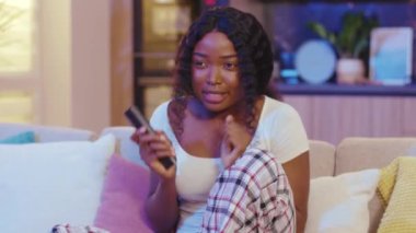 Siyah oturma odasındaki kanepede oturan Afrikalı Amerikalı kadın. Uzaktan kumandayla kanal değiştirmek geceleri televizyon izlemekten korkar. Yavaş çekim