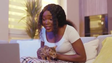 Kedi sohbeti yapan komik stil sahibi Afro-Amerikan kadın dizüstü bilgisayar kullanıyor. Evde video var. Mesafe ilişkisi. Gülümsüyor. Yavaş çekim