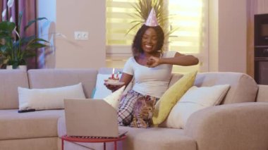 Çevrimiçi doğum günü partisini kediyle kutlayan Afrikalı Amerikalı bir kadın arkadaşlarıyla konuşurken pastadaki mumları üflüyor odada dizüstü bilgisayar kullanıyor. Evdeki kanepede tek başına oturuyorsun. Yavaş
