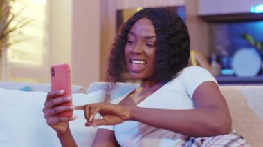 Portre Afro-Amerikan genç bayan rahat rahat koltuğa otur ve geceleri telefonu kullan. Rahat bir sosyal medya. Gülümseyen mutlu hisseder. Yavaş çekim