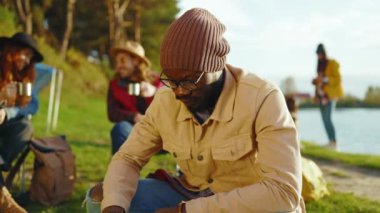 Tatlı Afro-Amerikan gözlüklü genç hippi kamp çadırında ateş yakıyor. Gülümseyen ve seyahat eden kameralara bakın. Göl kenarındaki ormanda. Kapatın. Yavaş çekim