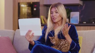 Sarışın, beyaz, güzel bir kadın. İnternetten arkadaşlarıyla tablet bilgisayarında video sohbeti yapıyor. Kanepede kedisini okşuyor. Sıradan bir yaşam tarzı. Teknoloji.