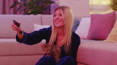 Bengal kedisiyle evde kalan genç bir kadın televizyon kanallarını değiştiriyor en sevdiği komik komedi dizisini seçiyor eğlencesine gülüyor. Evcil hayvan konsepti.