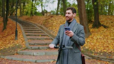 Sonbahar parkında genç ve şık bir adamın portresi elektrikli scooterlı telefon kulübesi kullanıyor. Ekolojik ulaşım. Gülümseyerek mutlu ol. Şehir dışından. Yavaş çekim