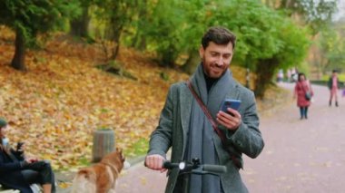 Autumn Park 'taki mutlu genç adam elektrikli scooterlı telefon kulübesi kullanıyor. Ekolojik ulaşım. Gülümsüyor. Şehir dışından. Yavaş çekim