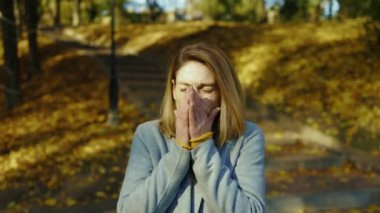 Güneş ışığında hasta genç bir kadın baş ağrısı çekerken hapşırıyor. Sonbahar parkında güneş ışığında kameraya bakıyor. Sarışın, stresli bayan. Portre ağrısı yorucu. Kapatın. Yavaş çekim