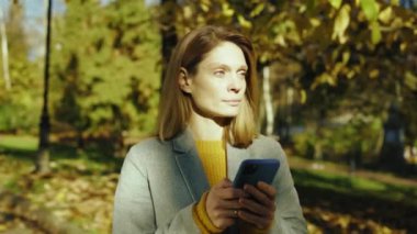 Palto standındaki genç kadın sonbahar parkında güneş ışığında hapşırıyor. Akıllı telefondan yazma teknolojisi. Portre. Kapatın. Yavaş hareket