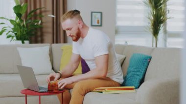 Çekici genç adam kanepede otururken dizüstü bilgisayar kullanıyor. Çay iç. Evde çalış. İnternet teknolojisi. Karantina zamanı. Kapatın. Yavaş çekim