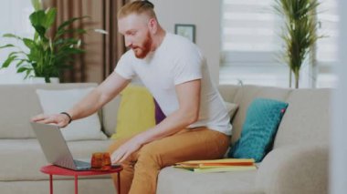 Mutlu genç adam kanepede otururken dizüstü bilgisayar kullanıyor. Çay iç. Evde çalış. İnternet teknolojisi. Karantina zamanı. Kapatın. Yavaş çekim