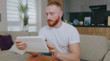 Tablet bilgisayar kullanan yakışıklı bir adam portresi. Odaklandım. Evdeki kanepede oturmak. Çevrimiçi teknoloji cihazı iletişim karantinası. Yavaş çekim