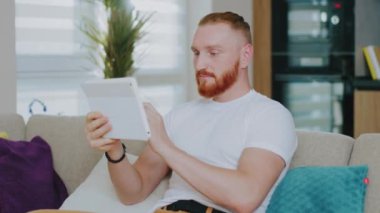 Tablet bilgisayar kullanan yakışıklı bir genç. Görüntülü konuşma. Evdeki kanepede oturmak. Çevrimiçi teknoloji aygıtı iletişim karantinası.