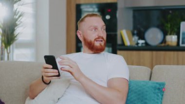 Çekici, yakışıklı, mutlu adam evinde, kanepede oturuyor. Akıllı telefon kullanarak dokunarak ve araklayarak. Sosyal medya interneti gülümsüyor. Bağlantı iletişimi. Kapat.