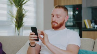 Evdeki yakışıklı, mutlu adam, akıllı telefon kullanırken koltuğa oturuyor, çeşitli dokunmalar yapıyor ve el kol hareketleri yapıyor. Rahat oturma odasında cep telefonuyla rahatlayan bir adam..