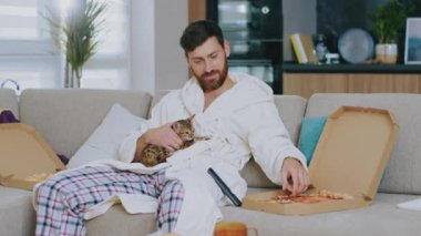 Kedili mutlu genç adam TV izlerken pizza yiyor. Evdeki kanepede oturmak. Dinlenme sinema kanalları dinleniyor. Portre. Kapatın. Yavaş çekim.