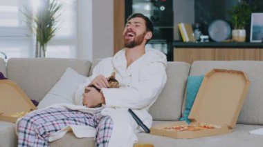 Kedili komik bir genç adam TV seyrediyor ve pizza yiyor. Kanepede oturup gülümsemek. Dinlenme sinema kanalları dinleniyor. Portre. Kapatın. Yavaş çekim.