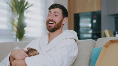 Pizza yiyen kedili genç bir adam portresi. Kanepede oturup gülümsemek. Dinlenme sinema kanalları dinleniyor. Portre. Kapatın. Yavaş çekim.