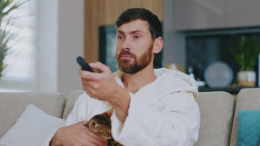 Kedili genç adam televizyon izliyor. Kanepede oturup gülümsemek. Dinlenme sinema kanalları dinleniyor. Portre. Kapatın. Yavaş çekim.