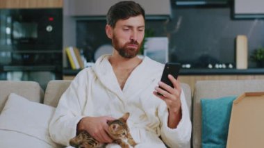 Kedili komik genç adam pizza yerken telefon kullanıyor. Kanepede oturup gülümsemek. Sosyal medyada dinlenme. Portre. Kapatın. Yavaş çekim.