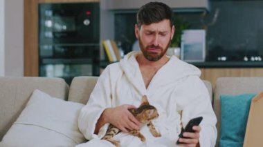 Kapat çeneni, kedisi olan hassas adam TV izlerken telefonu kullanıyor. Evdeki kanepede üzgün oturmak. Dinlenme sinema kanalları dinleniyor. Portre. Yavaş çekim.