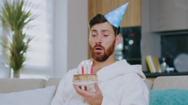Üzgün genç adam doğum gününü oturma odasında tek başına kutluyor karantina sırasında pastadaki mumları üflüyor ve televizyon seyrediyor. Kanepede oturmak. Covid-19. Evde kal. Depresyon. Yavaş çekim