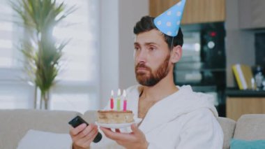 Portre genç adam oturma odasında yalnız başına doğum gününü kutluyor karantina sırasında pastadaki mumları üflüyor ve televizyon seyrediyor. Kanepede oturmak. Covid-19. Evde kal. Depresyon. Yavaş çekim