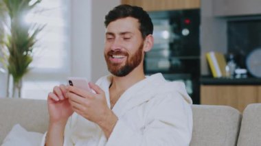 Yurt içi kıyafetleri içinde genç sakallı adam koltukta oturmuş gülümsüyor ve telefonu kullanıyor. Mutlu ol. Evde kal. Sosyal medya. İletişim konferansı portresi. Yavaş çekim