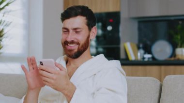 Gülümseyen, sakallı, yerli giysili çekici bir adam koltukta oturuyor ve telefon kullanıyor. Mutlu ol. Evde kal. Sosyal medya. İletişim konferansı portresi. Yavaş çekim