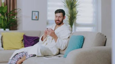Ev kıyafetleri içinde, kanepede oturmuş gülümseyen telefonu kullanan genç adam. Mutlu ol. Evde kal. Sosyal medya. İletişim konferansı portresi. Yavaş çekim