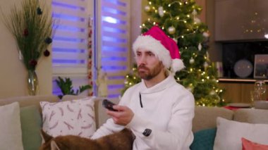 Ciddi, genç, şık bir adam, evinde köpek ile oturmuş, üzgün bir şekilde TV seyrediyor. Arka plandaki süslü Noel ağacında. Noel Baba kostümü. Yavaş çekim