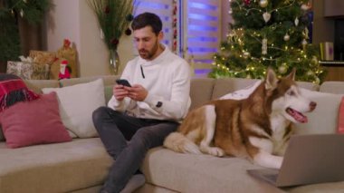 Evdeki kanepede köpekle oturan çekici genç adamın portresi. Telefonu kullan. Köpekle oyna. Arka plandaki süslü Noel ağacında. Yavaş çekim