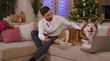 Dairede köpekle oturan çekici genç adamı kapat. Telefonu ve dizüstü bilgisayarı kullan. Köpekle oyna. Arka plandaki süslü Noel ağacında. Yavaş çekim