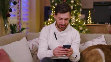 Gülümseyen genç adamın portresi evdeki kanepede oturmaktan mutluluk duyuyor. Noel zamanı. Yeni yıl kış tatili. Kutlama yapıyoruz. Mutlu noeller. Yavaş çekim