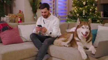 Mutlu, genç ve çekici bir adam evinde köpeğiyle oturuyor. Telefonla gülümsüyor. Köpekle oyna. Arka plandaki süslü Noel ağacında. Yavaş çekim