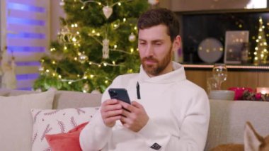 Evde gülümseyen genç adam koltukta oturmaktan mutlu olur. Noel zamanı. Yeni yıl kış tatili. Kutlama yapıyoruz. Mutlu noeller. Portre. Yavaş çekim