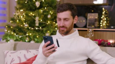 Gülümseyen genç adam, telefonu kullan. Evdeki kanepede oturmaktan mutlu ol. Noel zamanı. Yeni yıl kış tatili. Kutlama yapıyoruz. Mutlu noeller. Portre. Yavaş çekim