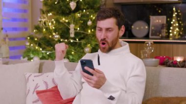 Gülümseyen mutlu genç adam evdeki koltukta oturan telefonu kullanıyor. Noel zamanı. Yeni yıl kış tatili. Kutlama yapıyoruz. Mutlu noeller. Portre. Yavaş çekim