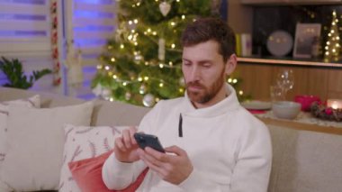 Üzgün genç adam evdeki kanepede otururken telefonu kullanıyor. Noel zamanı. Yeni yıl kış tatili. Kutlama yapıyoruz. Mutlu noeller. Portre. Yavaş çekim
