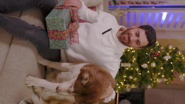 Çekici bir delikanlının arka plandaki Noel süslemelerinde köpek tutacağı hediyesiyle kanepede oturduğu dikey çekimini kapat. Mutlu bir hayvan portresi. Noel zamanı. Yavaş çekim