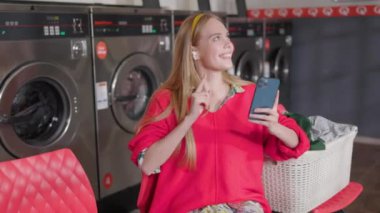 Güzel sarışın kadın telefon ve kulaklık kullanıyor. Çamaşır odasında oturmuş çamaşır yıkıyor. Ev işi çamaşır makinesi kurutma makinesi içeride. Kapatın. Yavaş çekim